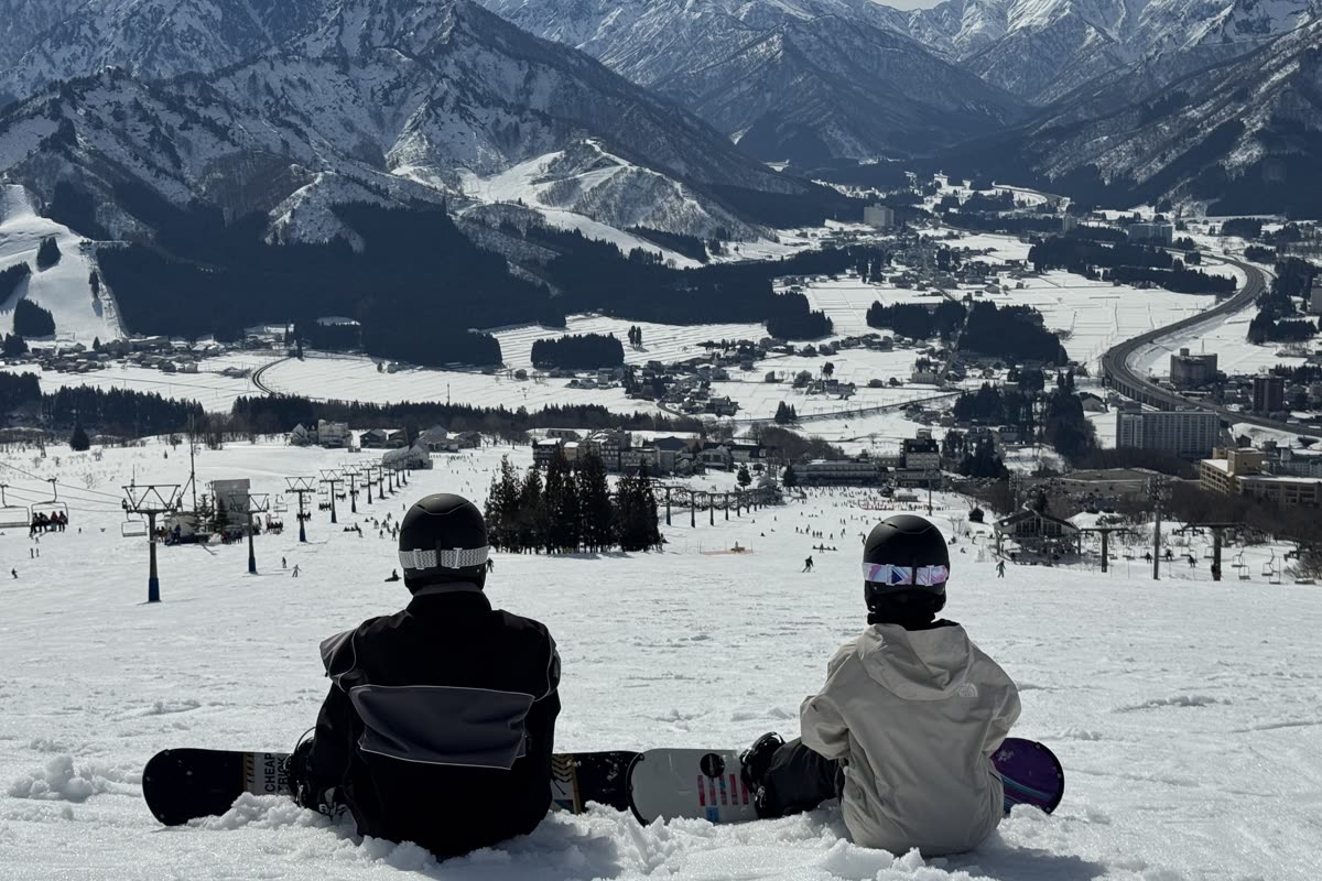 兩位單板玩家並肩坐在雪地上,背對鏡頭眺望遠方城鎮與雪山
