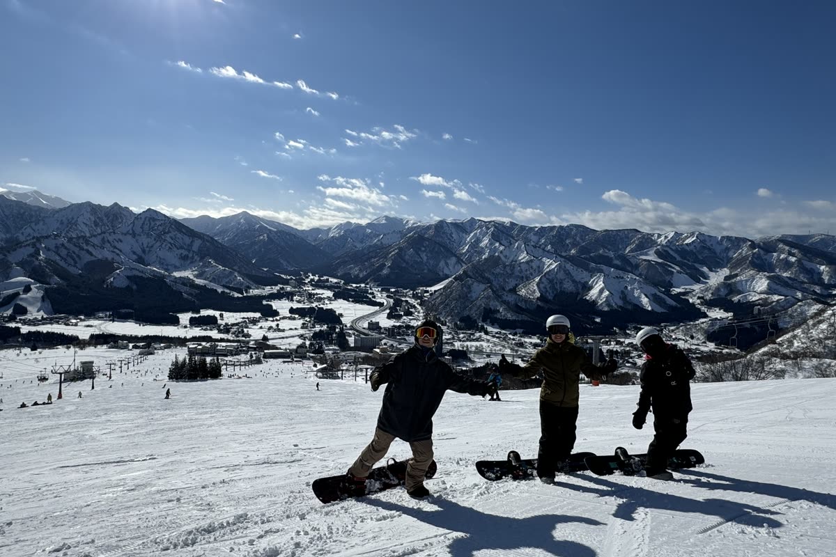 三位單板玩家站在岩原的雪道頂端張開雙手,背景是湯澤市街與群山